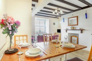 A dining area with a wooden table set for four, a vase of flowers, and a view of the living room in the background. - 3 Bed in Aberdaron (oc-wag407) (Aberdaron)