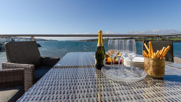 A table set with a bottle of champagne, glasses, plates, and a basket of baguettes on a deck with a view of the water.
