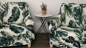 Två fåtöljer med ett tropiskt bladmotiv, ett litet runt bord med en krukväxt och ett fönster med utsikt över blommor.