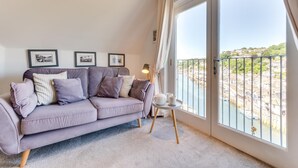 Un salón moderno con un sofá gris, una mesita redonda con una lámpara y un ventanal con vistas a un río y edificios.