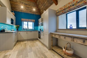 Eine Küche mit blauer Wand, Holzbalkendecke, einem Fenster mit floralem Vorhang und einer Holzbank mit Korb.