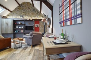 Ein gemütliches Wohnzimmer mit Stein Decke, Kamin, Essbereich mit Tisch für zwei Personen und Fernseher auf einem Regal.