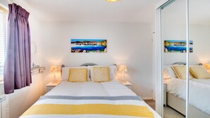 A bedroom with a bed, two bedside tables, a nightstand, a mirror, and a painting of a coastal scene. - 3 Bed in Abersoch (oc-kook72) (Abersoch)