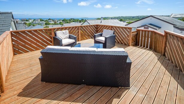 A wooden deck with wicker furniture, including a sofa and two chairs, overlooking a scenic view of houses and the sea. - 3 Bed in Abersoch (oc-kook72) (Abersoch)