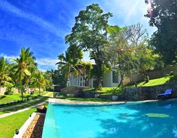 Una piscina con muro de piedra y palmeras, rodeada de césped cuidado y un edificio con columnas.