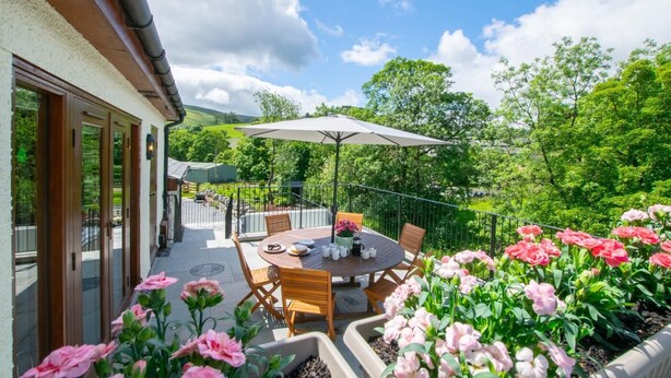 Een terras met een ronde tafel, stoelen en een grote parasol. Rond de tafel staan bloempotten en bloemen.
