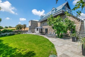 A modern house with a stone exterior, a patio with outdoor furniture, and a clear blue sky. - 5 Bed in Little Petherick (oc-p00736) (Tregonna)