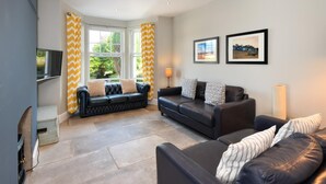 A modern living room with a black leather sofa, a smaller sofa with patterned cushions, and a large window with yellow curtains. There are two framed pictures on the wall and a lamp on the floor.