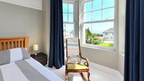 A bedroom with a bed, a nightstand, a chair, and large windows with a view of houses and greenery.