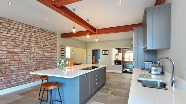 A modern kitchen with a central island, bar stools, a brick accent wall, and a view into the living area.
