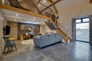 A spacious living area with a grey sofa, a wooden staircase, and a stone wall. - 2 Bed in Aberdaron (oc-bow86) (Rhoshirwaun)