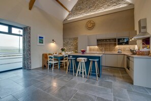 A modern kitchen with a stone accent wall, a clock, and a dining area with stools. - 2 Bed in Aberdaron (oc-bow86) (Rhoshirwaun)