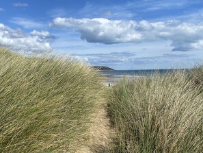 Uma trilha de areia ladeada por grama alta que leva a uma praia, com céu limpo e colinas ao longe.
