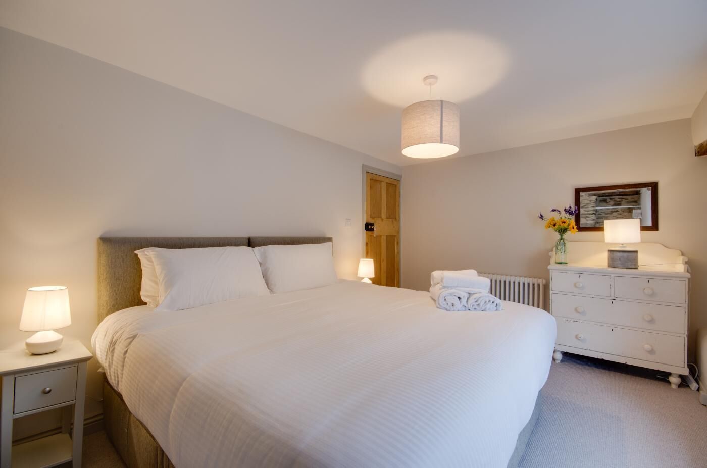 Un lit bien fait, avec du linge de lit blanc, une lampe de chevet, une commode avec un miroir et un radiateur.