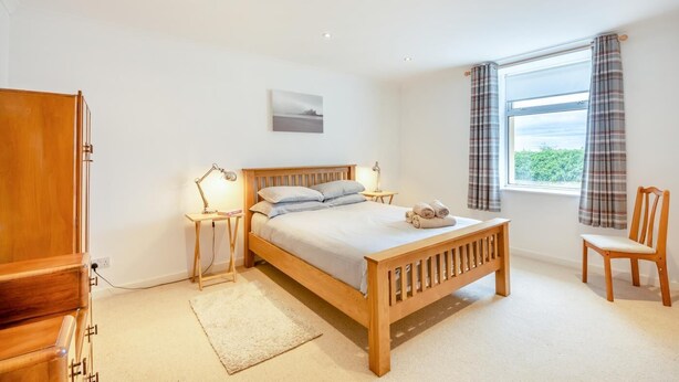 A bedroom with a wooden bed, bedside lamps, a chair, a window with curtains, and a view of greenery outside. - 2 Bed in Lowick (oc-414296) (Lowick)