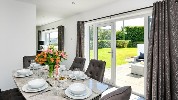 Un coin repas avec une table en marbre dressée pour quatre personnes, entourée de chaises, et une vue sur un jardin grâce à de grandes portes-fenêtres coulissantes.