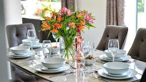 Une table à manger dressée avec de l’assiette blanche, des bols et des verres à vin, agrémentée d’un vase de fleurs.