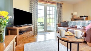 Uma sala de estar com televisão, sofá, mesa de centro com xícaras e doces, e vista através de uma porta de vidro.