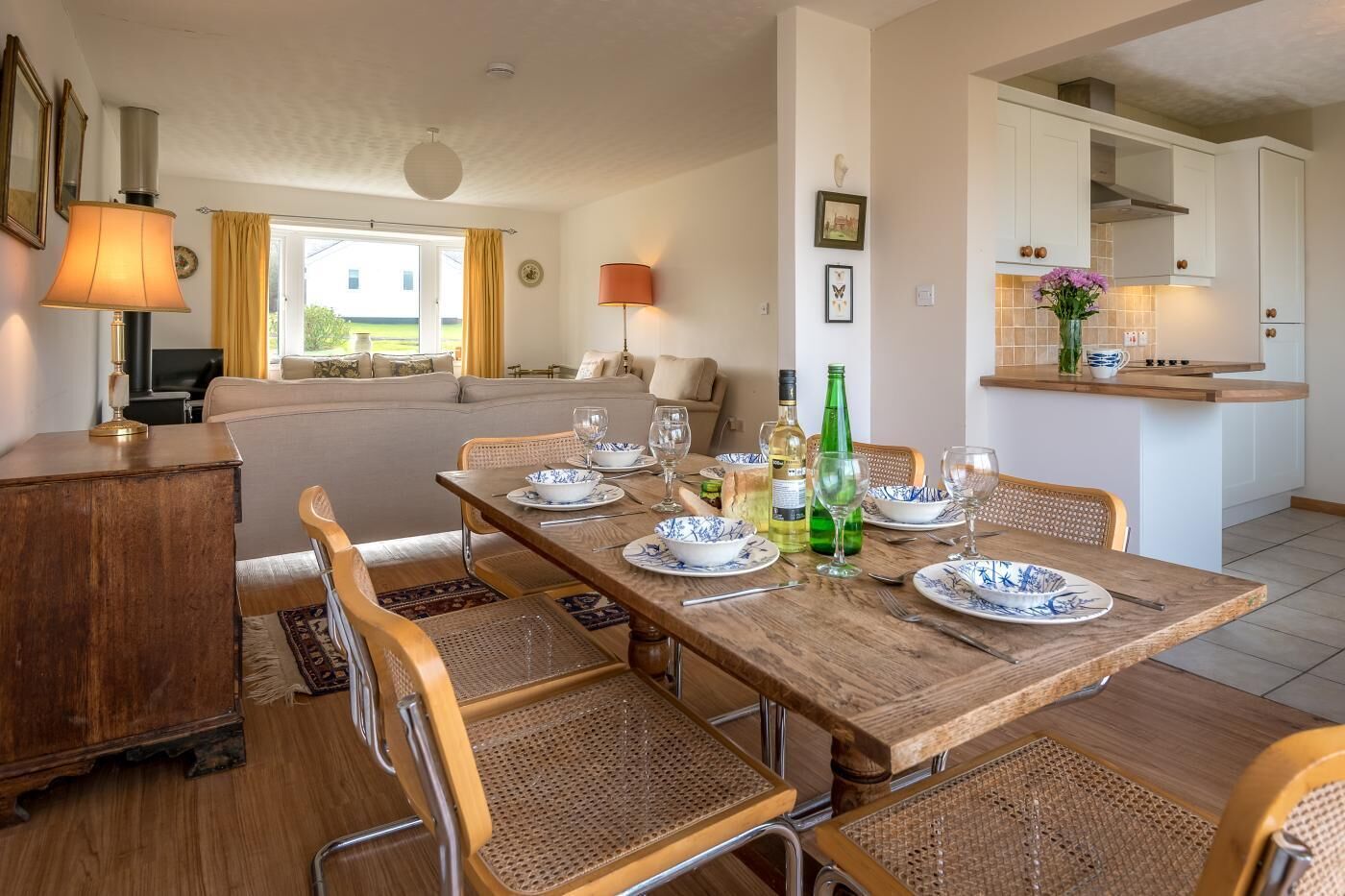 A dining area with a wooden table set for six, surrounded by wicker chairs. A kitchen with white cabinets and a countertop is visible in the background.