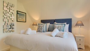 Een net opgemaakt bed met witte lakens en kussens, een aan de muur bevestigd abstract kunstwerk en twee bedlampen op een nachtkastje.