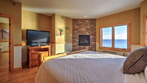 Een slaapkamer met een groot bed, een televisie op een houten bureau, een stenen muur en een raam met uitzicht op zee.