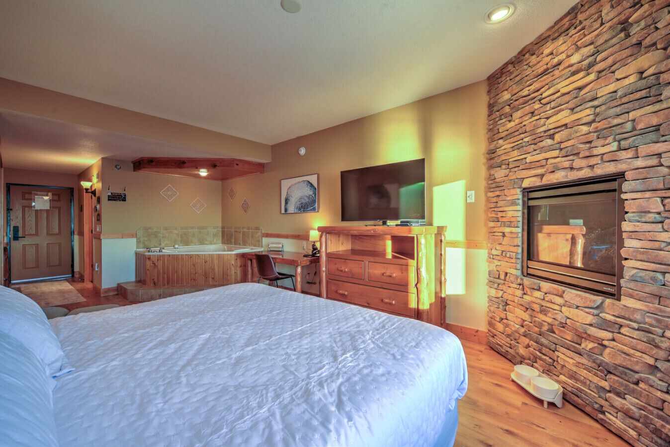 Ein Hotelzimmer mit einem großen Bett, einem Holztisch, einem Fernseher, einer Steinwand und einem Badezimmer mit Badewanne und Waschbecken.
