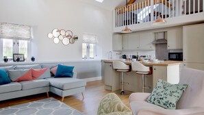 A spacious living area with a grey sofa, a pink armchair, and a small table. A kitchen with a bar area is visible in the background.