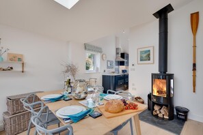 A modern kitchen with a wood-burning stove, dining table set for four, and a view of the outdoors. - 2 Bed in Polruan (oc-p00896) (England)