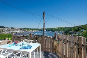 An outdoor dining area with a view of a harbor, wooden fence, and a table set with plates and a bowl of fruit. - 2 Bed in Polruan (oc-p00896) (England)