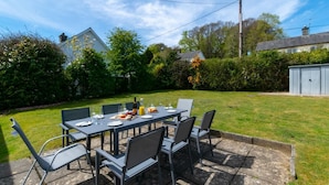 Eine Terrasse mit Tisch für vier Personen, umgeben von Stühlen, mit einer Rasenfläche und einem Schuppen im Hintergrund.