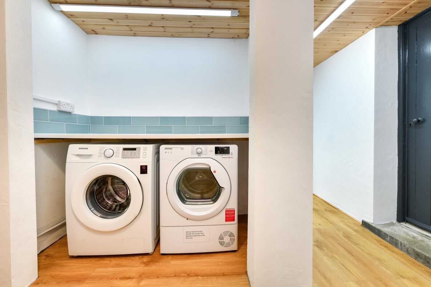 A laundry room with two white front-loading washing machines, a wooden ceiling, and a blue tiled backsplash.