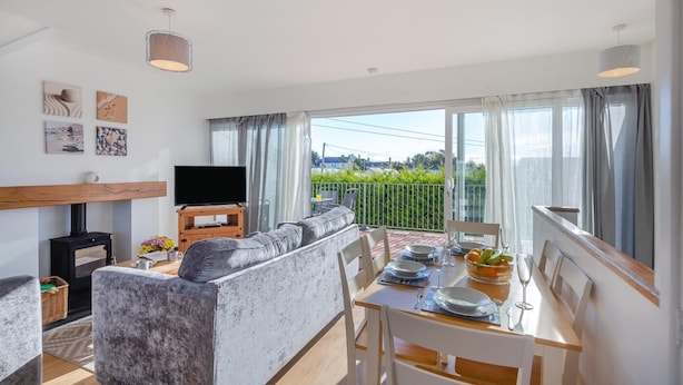 Ein modernes Wohnzimmer mit einem grauen Sofa, einem Holz-TV-Schrank, einem Kamin und einem Essbereich mit Tisch für zwei Personen.