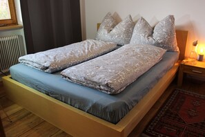 Een bed met een gebedrukte deken en kussens, een houten hoofdbord, een nachtkastje met een lamp, en een raam met gordijnen.