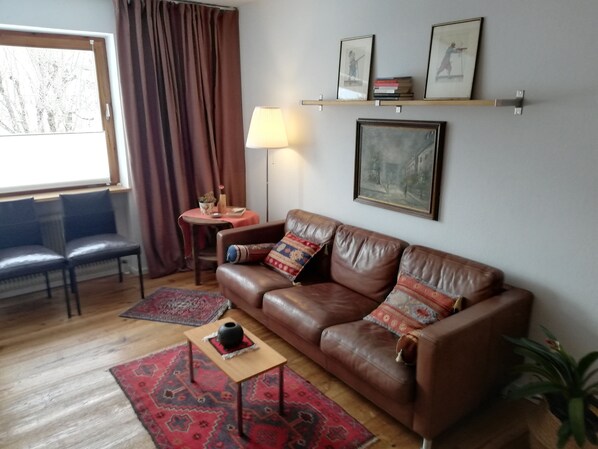 Een woonkamer met een bruine leren bank, twee fauteuils, een klein tafeltje met een lamp, een rood tapijt met dessin en een potplant.