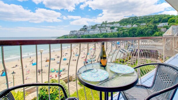 A balcony with a table and chairs overlooking a beach with umbrellas and people. - 3 Bed in Looe (oc-5adm) (Looe)