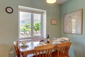 A dining table with chairs, a wine bottle, and bowls set for a meal. A window with a view of greenery and mountains is visible. - 2 Bed in Nant Peris (oc-wag693) (Nant Peris)