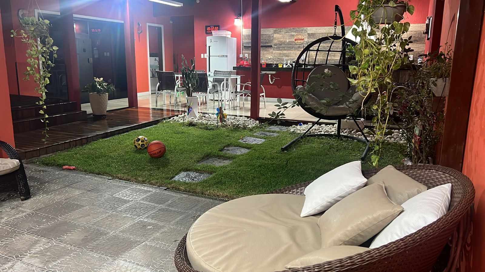 A cozy outdoor seating area with a wicker chair and cushions, a small lawn with a soccer ball, and a dining area with chairs and a table.