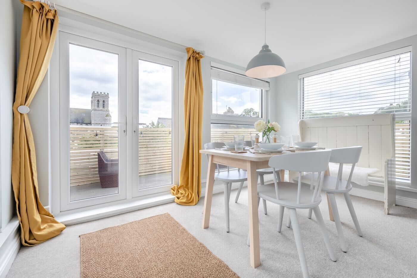 A dining area with a table set for two, white chairs, a grey pendant light, and a view through a glass door.