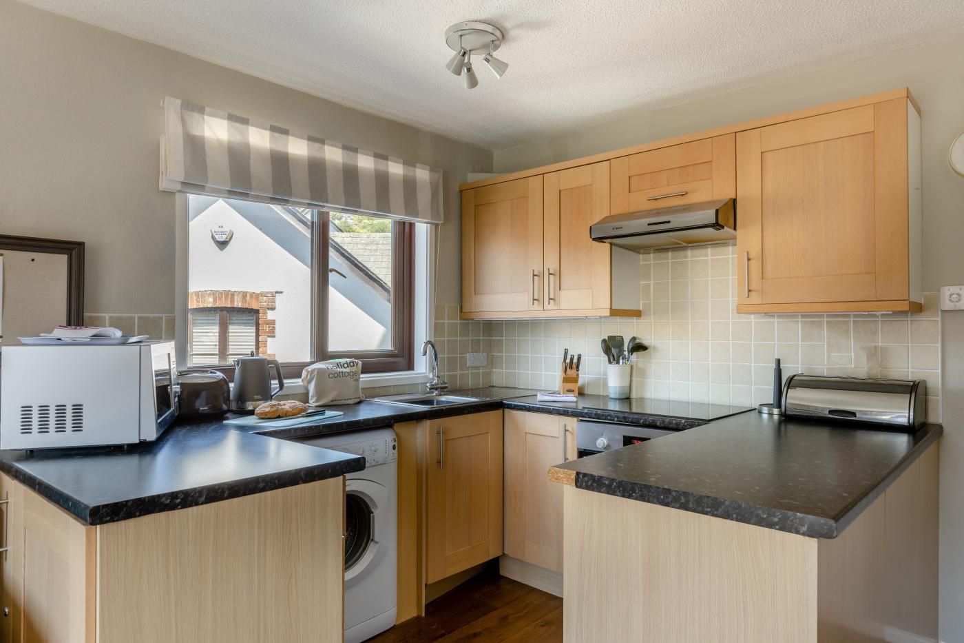 A compact kitchen with wooden cabinets, a built-in washing machine, a toaster, and a window with a view of the outside.