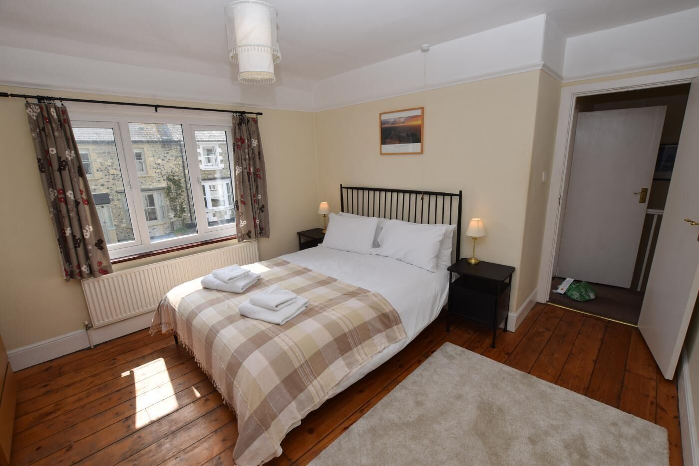 A bedroom with a bed, two bedside tables, a window with curtains, a radiator, a door, and a small rug.
