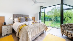 Une chambre moderne avec un grand lit, des tables de chevet, un ventilateur de plafond et une vue sur un jardin grâce à de grandes fenêtres.