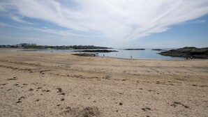 Uma extensa praia de areia clara, com águas calmas, formações rochosas e edifícios ao longe.