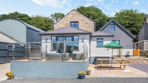 A modern house with a stone roof, a glass enclosure with wicker furniture, and a picnic table with an umbrella. - 3 Bed in Talwrn (oc-bow229) (Ysgubor Hen)