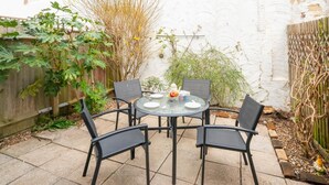 Et rundt glasbord med fire stole, placeret på en terrasse med en hvid væg og planter.