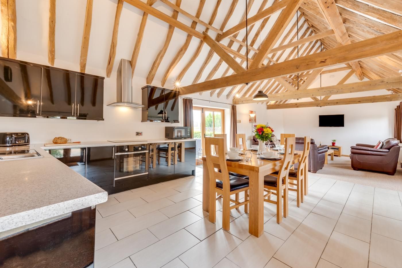 A spacious kitchen with wooden beams, a dining area with chairs, and a living room with a sofa and TV.