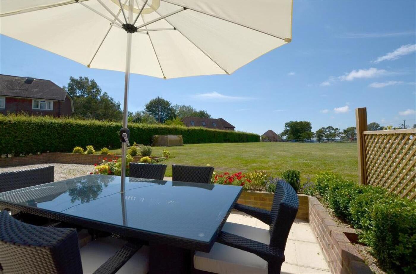 A patio with a glass table and wicker chairs, surrounded by a hedge and a wooden fence.