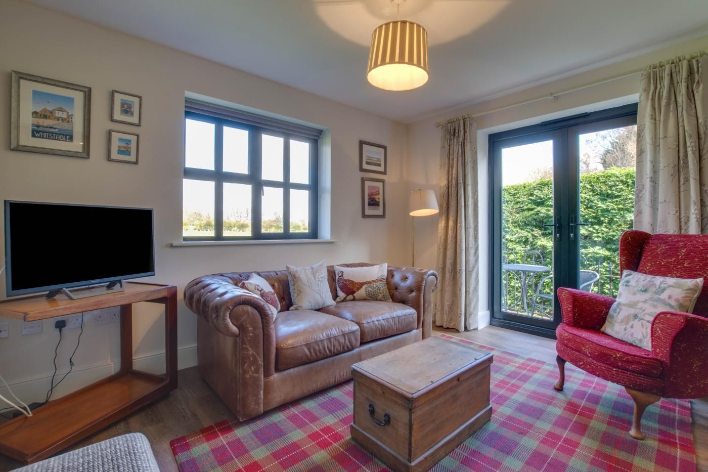A living room with a leather sofa, a red armchair, a wooden coffee table, a flat-screen TV on a wooden stand, and a large window with curtains.
