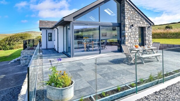 A modern house with a glass conservatory, stone exterior, and a patio with outdoor furniture.