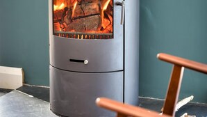A modern wood-burning stove with visible flames, set against a teal wall, in a room with a tiled floor and wooden furniture.
