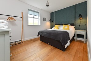 Une chambre à coucher avec un lit, un plancher en bois, une fenêtre avec des stores, une étagère fixée au mur et une table de chevet avec une lampe.